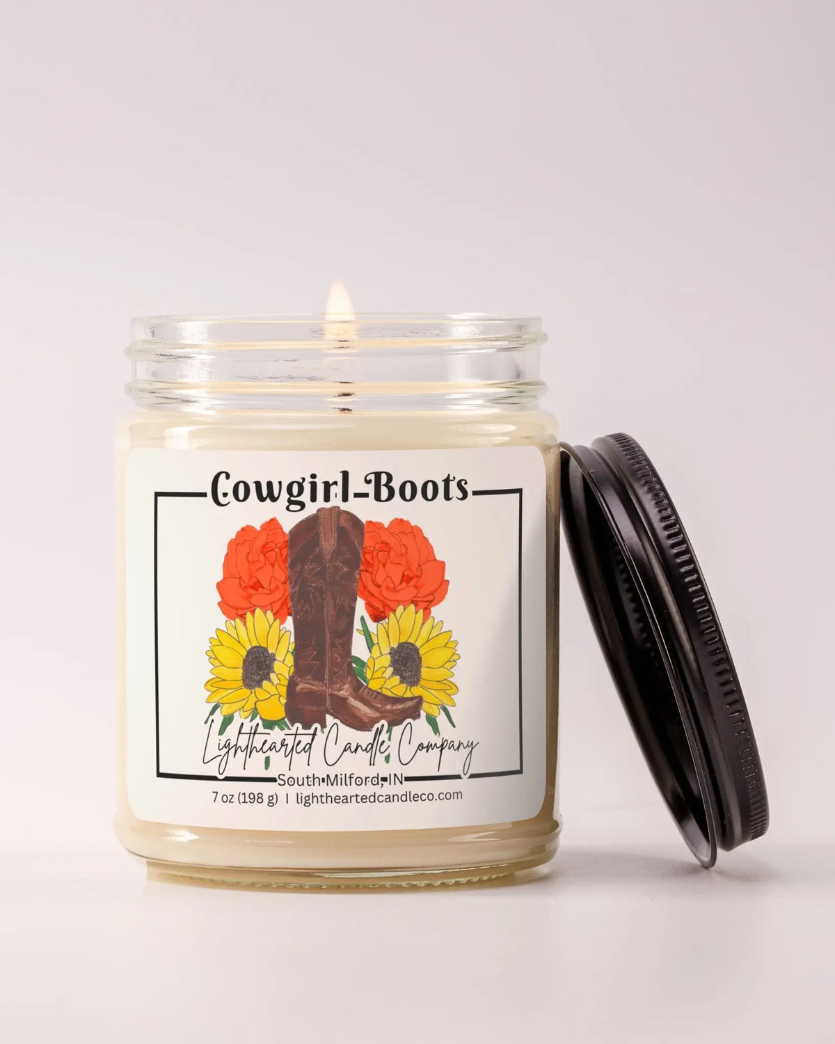 Lit Cowgirl Boots Candle in a clear glass jar with its black lid beside it