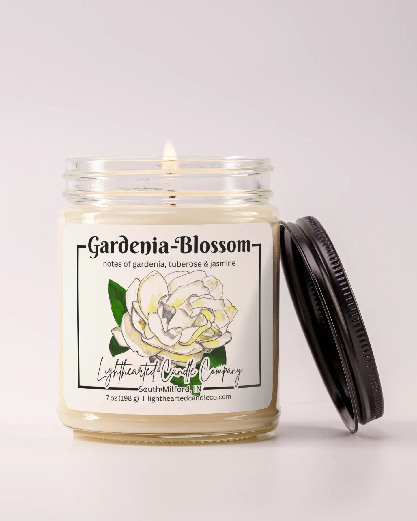 Lit Gardenia Blossom Candle in a clear glass jar with its black lid beside it