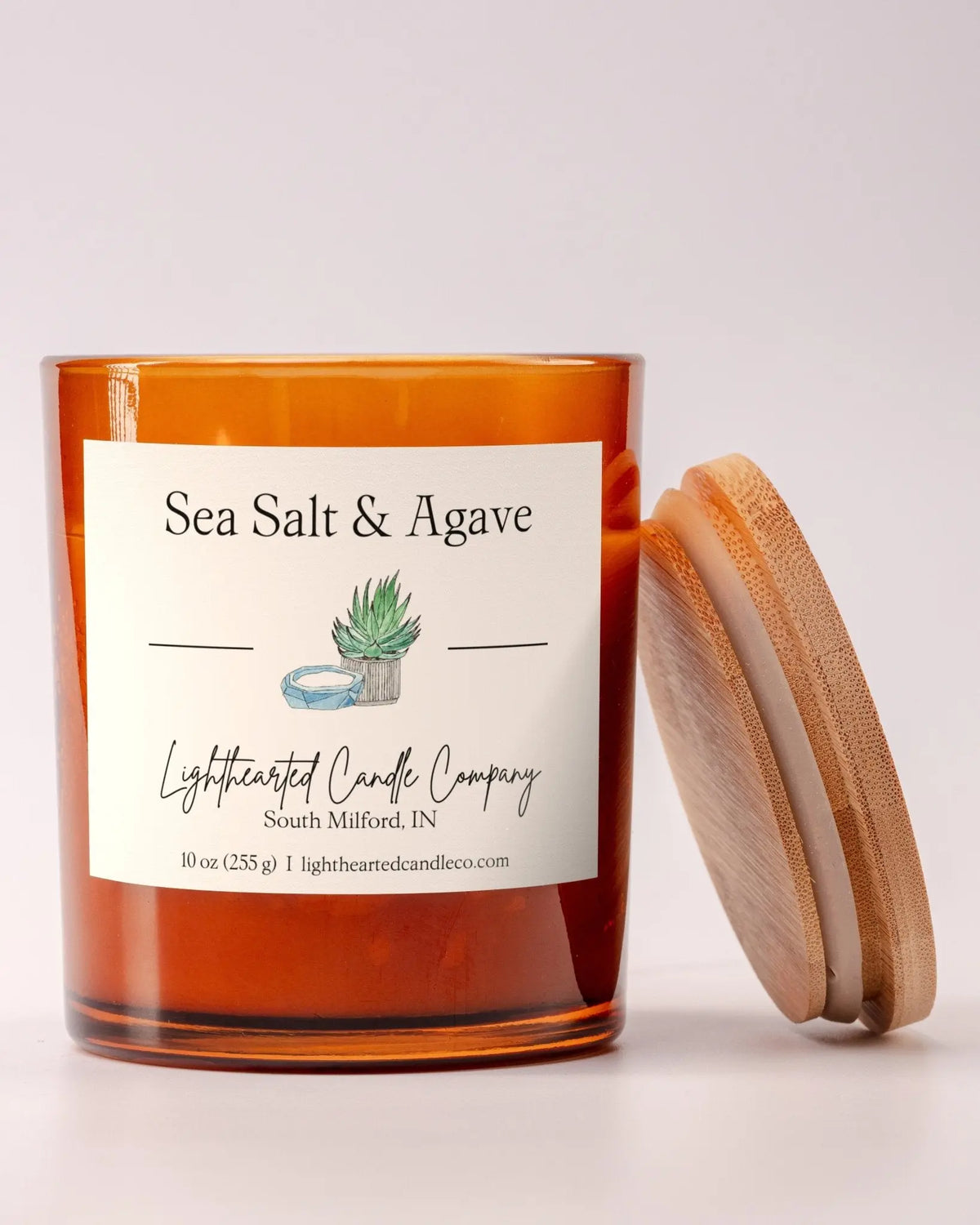 Lit Sea Salt & Agave Candle in an amber glass jar with its wooden lid leaning beside it