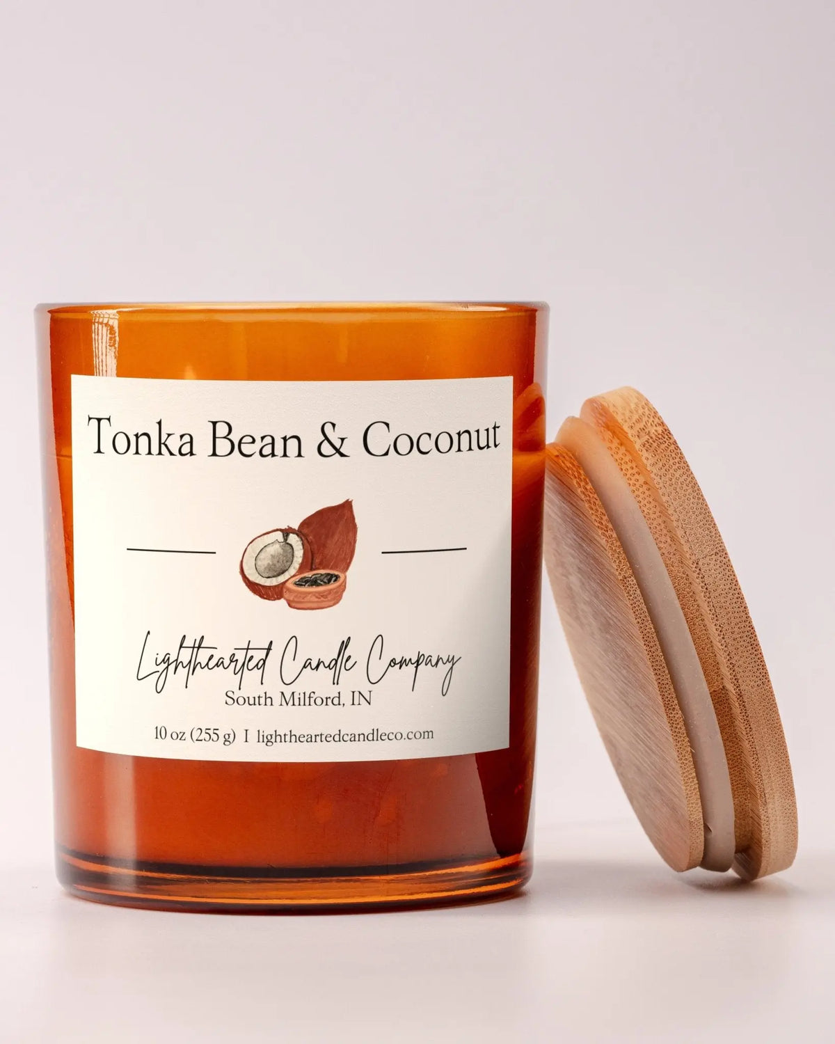 Lit Tonka Bean & Coconut Candle in an amber glass jar with its wooden lid leaning beside it