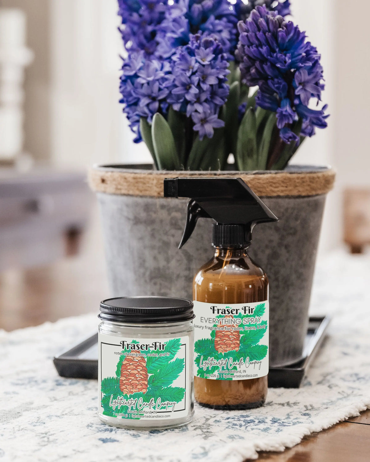 Matching candle and Fraser Fir Everything Spray styled beside purple flowers