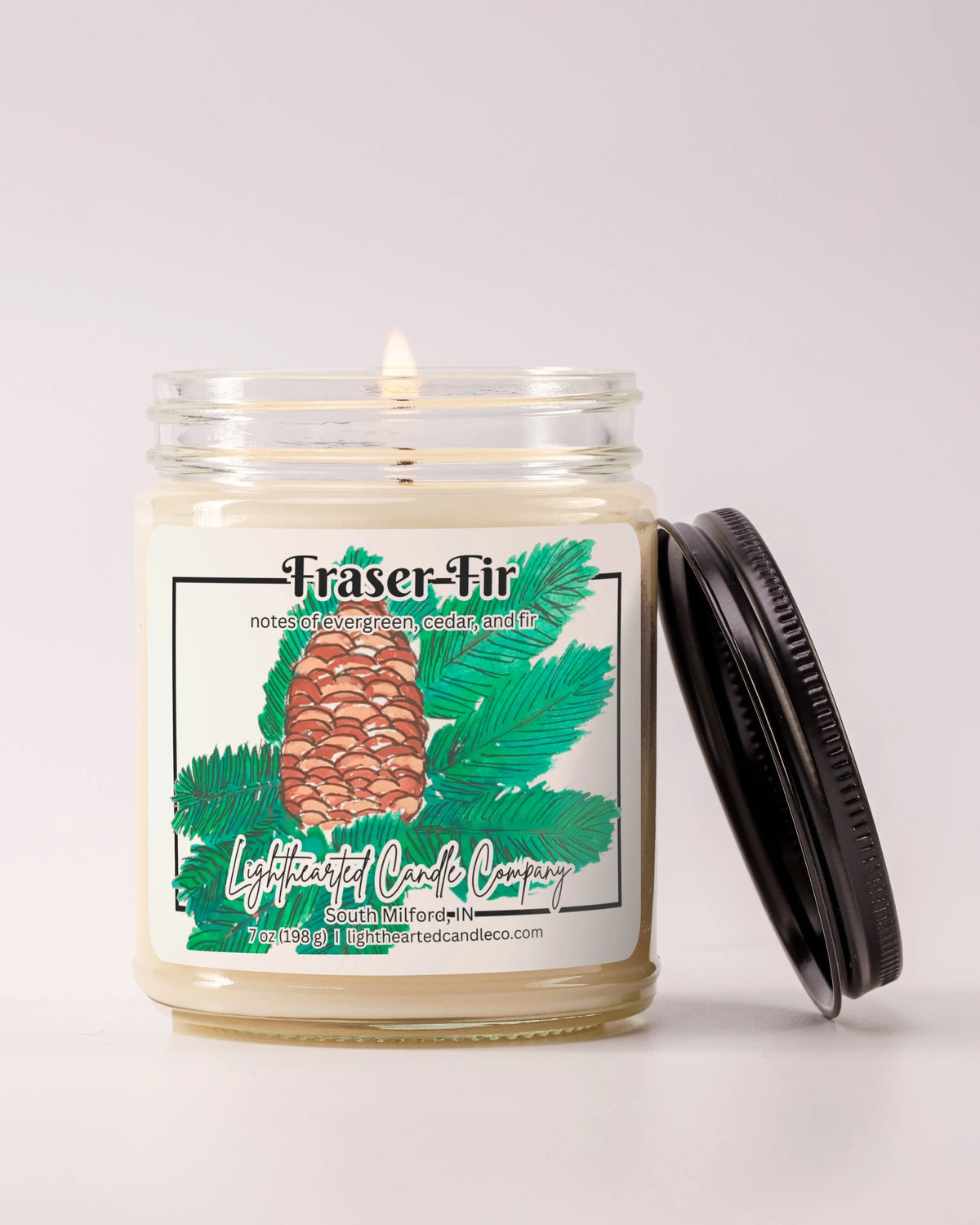 Lit Fraser Fir Candle in a clear glass jar with its black lid beside it