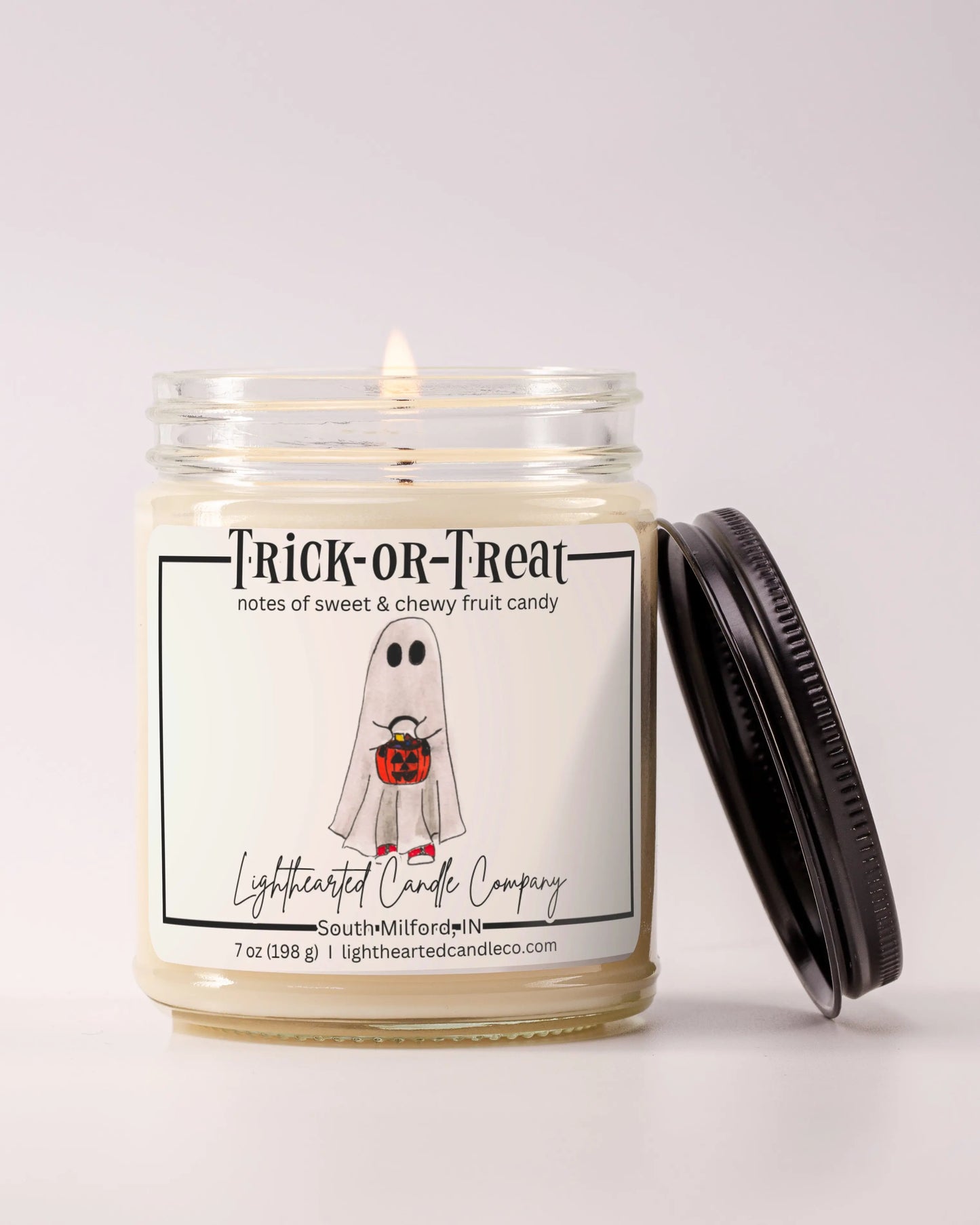 Lit Trick or Treat Candle in a clear glass jar with its black lid beside it, alternate view 6