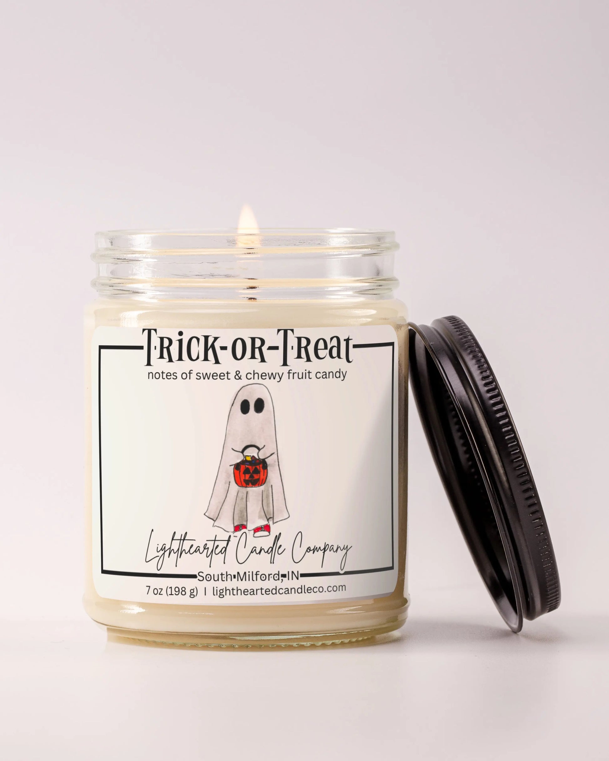 Lit Trick or Treat Candle in a clear glass jar with its black lid beside it, alternate view 6