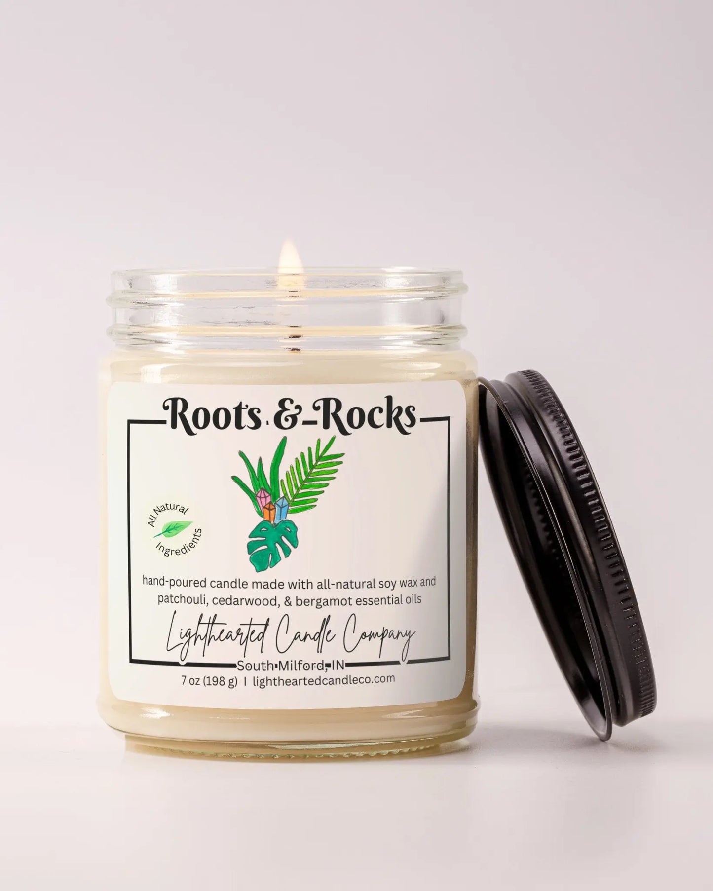 Lit Roots & Rocks Essential Oil Candle in a clear glass jar with its black lid beside it