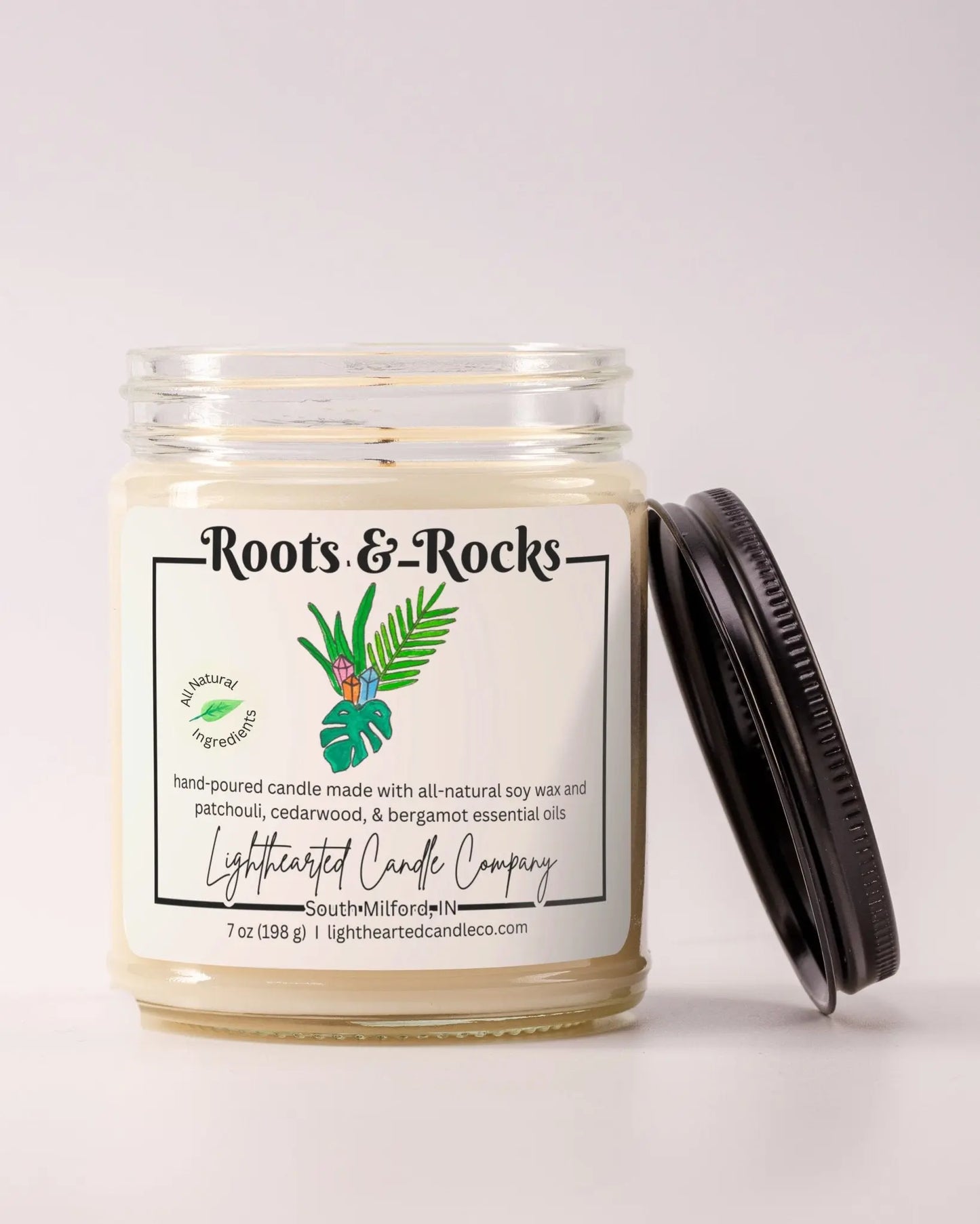 Unlit Roots & Rocks Essential Oil Candle in a clear glass jar with its black lid beside it